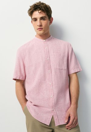 Jeune homme aux cheveux courts et bouclés portant une chemise rose clair à manches courtes et un pantalon beige, debout devant un fond blanc.