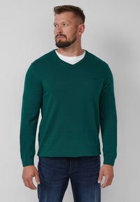 Grüner Strickpullover mit V-Ausschnitt, langen Ärmeln und gerippten Bündchen. Getragen über einem weißen Hemd, kombiniert mit blauen Jeans.