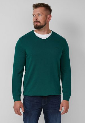 Grüner Strickpullover mit V-Ausschnitt, langen Ärmeln und gerippten Bündchen. Getragen über einem weißen Hemd, kombiniert mit blauen Jeans.