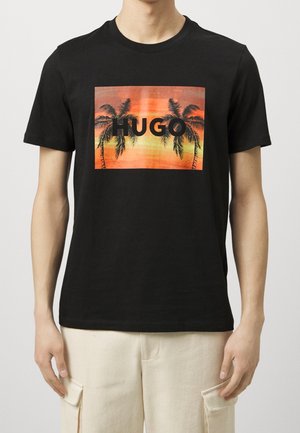 T-shirt en coton noir avec un graphique sur le devant représentant des palmiers sur fond de coucher de soleil orange, avec le mot "HUGO" en lettres noires en gras.
