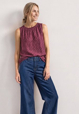 Femme aux cheveux blonds attachés, portant un haut bordeaux texturé sans manches et un jean bleu large, debout contre un mur clair.
