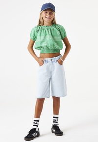 Top corto verde con estampado y cintura elástica, combinado con pantalones cortos de mezclilla azul claro. Zapatillas negras y calcetines blancos con detalles negros.