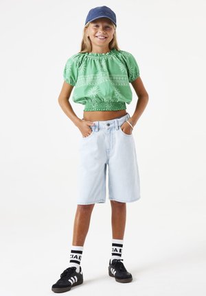 Croptop vert à motifs avec taille élastique, associé à un short en denim bleu clair. Baskets noires et chaussettes blanches avec accents noirs.