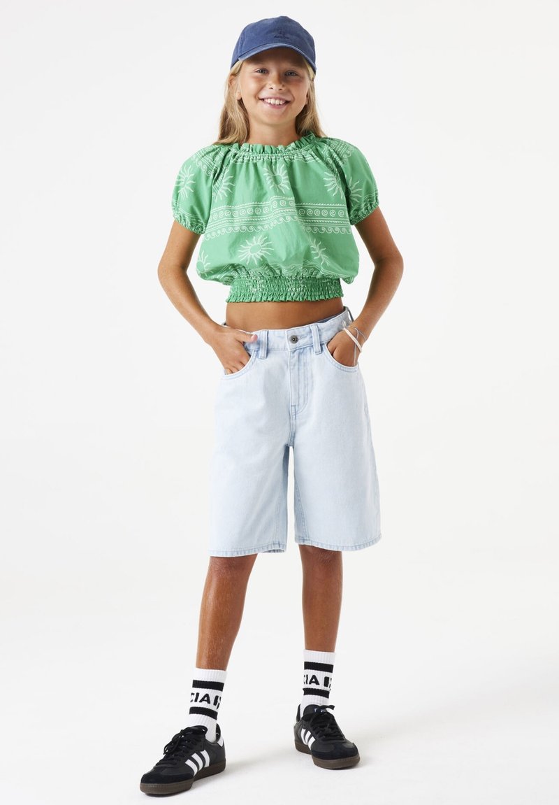 Top corto verde con estampado y cintura elástica, combinado con pantalones cortos de mezclilla azul claro. Zapatillas negras y calcetines blancos con detalles negros.