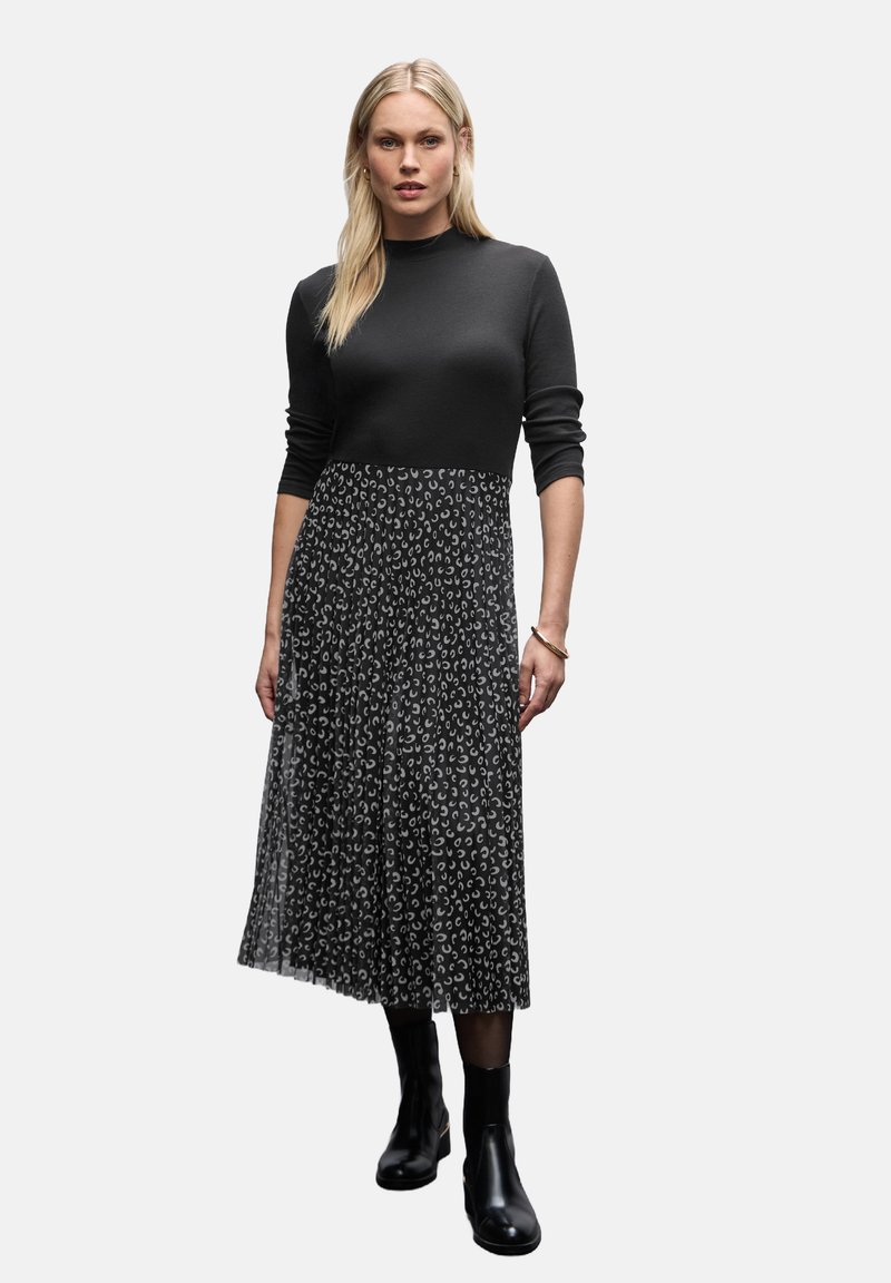 Vrouw met lang blond haar, gekleed in een zwart langslachtige top, een gemusterde plissé midi rok, zwarte panty's en zwarte enkellaarsjes, rechtopstaand.
