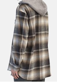 Rutigt skjorta i brunt, svart och krämfärgat med en grå huvtröja. Har långa ärmar, en avslappnad passform och knäppningar vid manschetterna på ett strukturerat tyg.