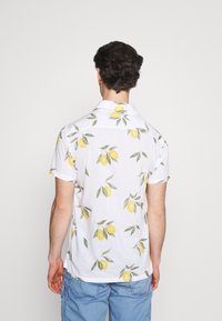 Man met krullend donker haar, gekleed in een witte short-sleeve shirt met een patroon van gele citroenen en groene bladeren, gecombineerd met lichtblauwe cargoshorts, met zijn rug naar ons toegekeerd.