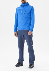Homme portant une veste zippée bleue avec des poches sur la poitrine et sur les côtés, un pantalon bleu foncé avec des poches zippées sur les cuisses, et des chaussures de randonnée noires.