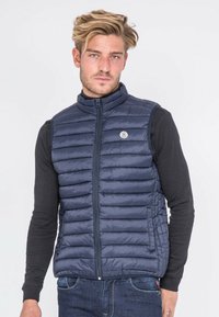 Veste matelassée marine avec des compartiments verticaux, col haut et fermeture éclair intégrale. Présente un badge logo et est portée par-dessus une chemise noire à manches longues.