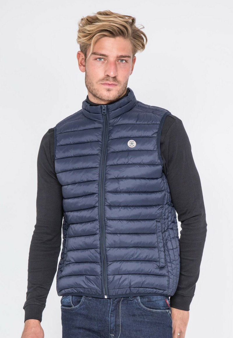 Veste matelassée marine avec des compartiments verticaux, col haut et fermeture éclair intégrale. Présente un badge logo et est portée par-dessus une chemise noire à manches longues.