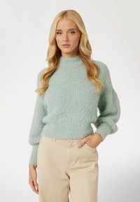 Minkštas, pūkuotas šviesiai žalias megztinis; turi aukštą iškirpę, laisvą siluetą ir balioninius rankovės. Suderintas su šviesiai beigės spalvos kelnėmis.