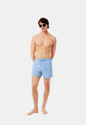 Hemdloser Mann trägt hellblaue Badeshorts und Sonnenbrille und steht barfuß vor einem schlichten hellen Hintergrund.