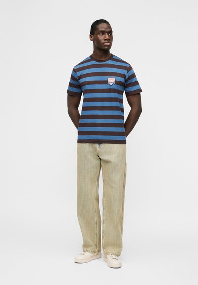 Άνδρας, όρθιος με τα χέρια στην πλάτη, φοράει μπλούζα με μπλε και καφέ ρίγες, ανοιχτό μπεζ τζιν με φαρδιά πόδια και μπεζ αθλητικά παπούτσια.