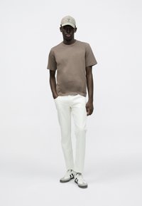 Καφέ βαμβακερό μπλουζάκι, λευκό τζιν παντελόνι, γκρι και μαύρα ριγέ αθλητικά παπούτσια, γαλάζιο καπέλο. Το μοντέλο στέκεται με το ένα χέρι στην τσέπη.