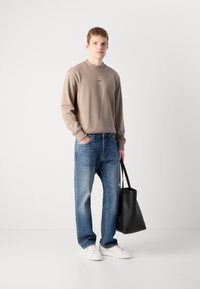 Joven vestido con sudadera beige, jeans azules, zapatillas blancas, sosteniendo una bolsa tote negra frente a un fondo blanco liso.