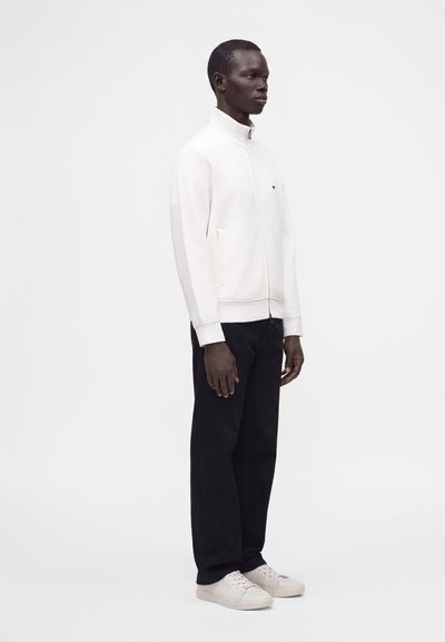 Homme portant une veste blanche à fermeture éclair, un pantalon noir et des baskets blanches, debout de côté sur un fond blanc uni.