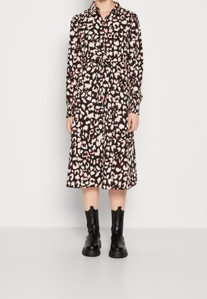 Vrouw die een midi-jurk draagt met lange mouwen, knopen en een ceintuur, met een luipaardprint in zwart, wit en donkerrood, gecombineerd met zwarte enkellaarzen.
