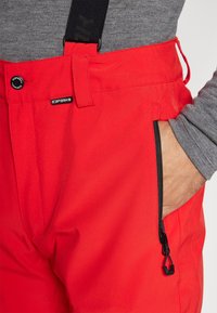 Pantalon de ski rouge avec bretelles noires réglables, doté de poches zippées et d'un tissu lisse et résistant à l'eau. Logo visible sur la ceinture.