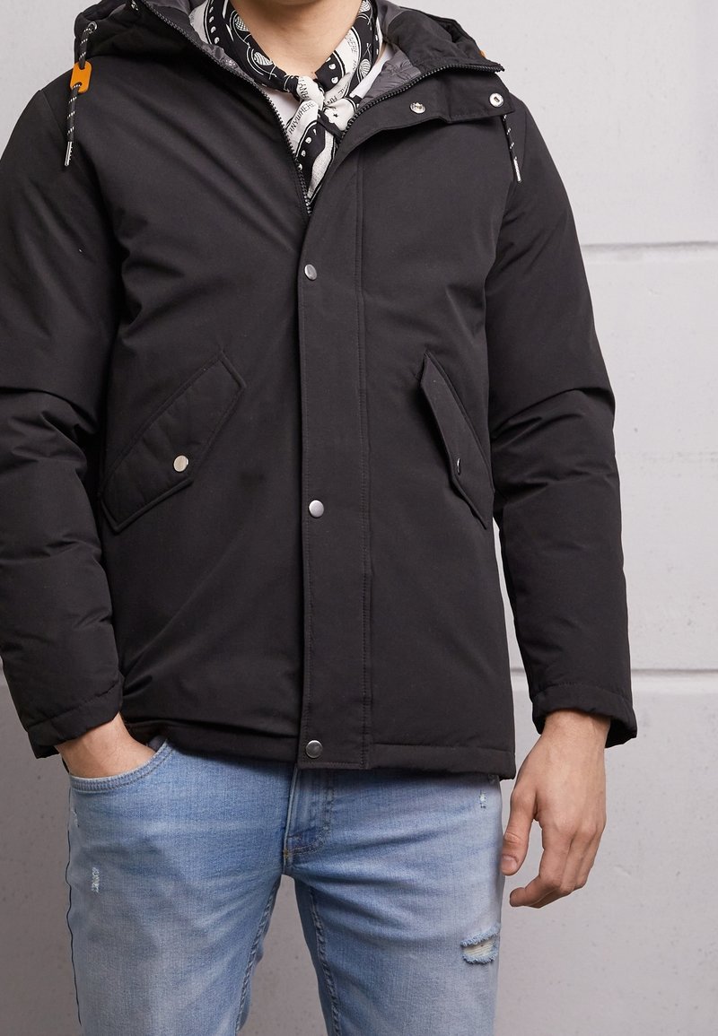 Svart vadderad jacka med huva, tryckknappar och två framfickor. Har ett innerfoder med en mönstrad scarf synlig vid halsen.