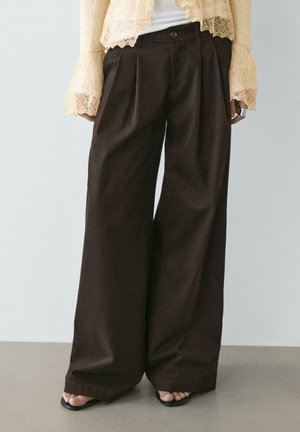 Pantalon noir à jambes larges avec plis devant, porté avec des manches en dentelle beige et des sandales noires à bout carré laissant apparaître les orteils.