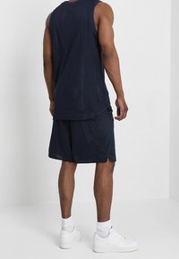 Man står med ryggen mot kameran, iförd en marinblå ärmlös meshtröja, matchande shorts, vita strumpor och vita sneakers mot en enfärgad bakgrund.