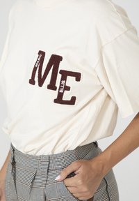 Crème kleurige t-shirt met korte mouwen met gestructureerde bruine "ME" letters. Gecombineerd met hoog-getailleerde geruite broeken die een getailleerde pasvorm tonen.