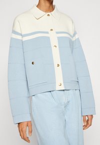 Jasnoniebieska krótka kurtka z białym kołnierzem i poziomymi paskami. Posiada złote guziki i boczne kieszenie; teksturowana tkanina.