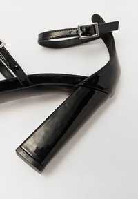 Sandale haute en cuir verni noir à talon bloc, avec une bride de cheville ajustable et une texture lisse et élégante.