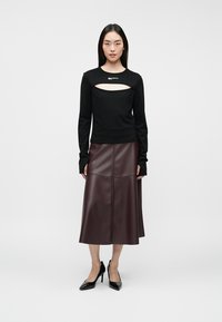 Sieviete, ģērbusies melnā garajā piedurknē t-kreklā ar krūšu izgriezumu, bordo ādas midi svārkos un melnās norādītāju kurpēs, stāv uz vienkāršas fona.