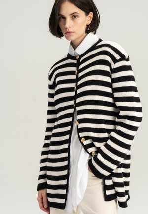 Black and white striped cardigan with a relaxed fit, knit texture, gold buttons, and a round neck, layered over a white shirt.