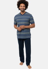 Man met krullend haar glimlacht, gekleed in een blauw gestreept T-shirt met korte mouwen en marineblauwe pyjamabroek, staande op blote voeten voor een effen achtergrond.
