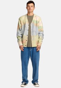 Gebreide cardigan met pastel abstracte patronen, lichte kleuren en een ruime pasvorm, gecombineerd met losse denim broeken en zwarte sneakers.