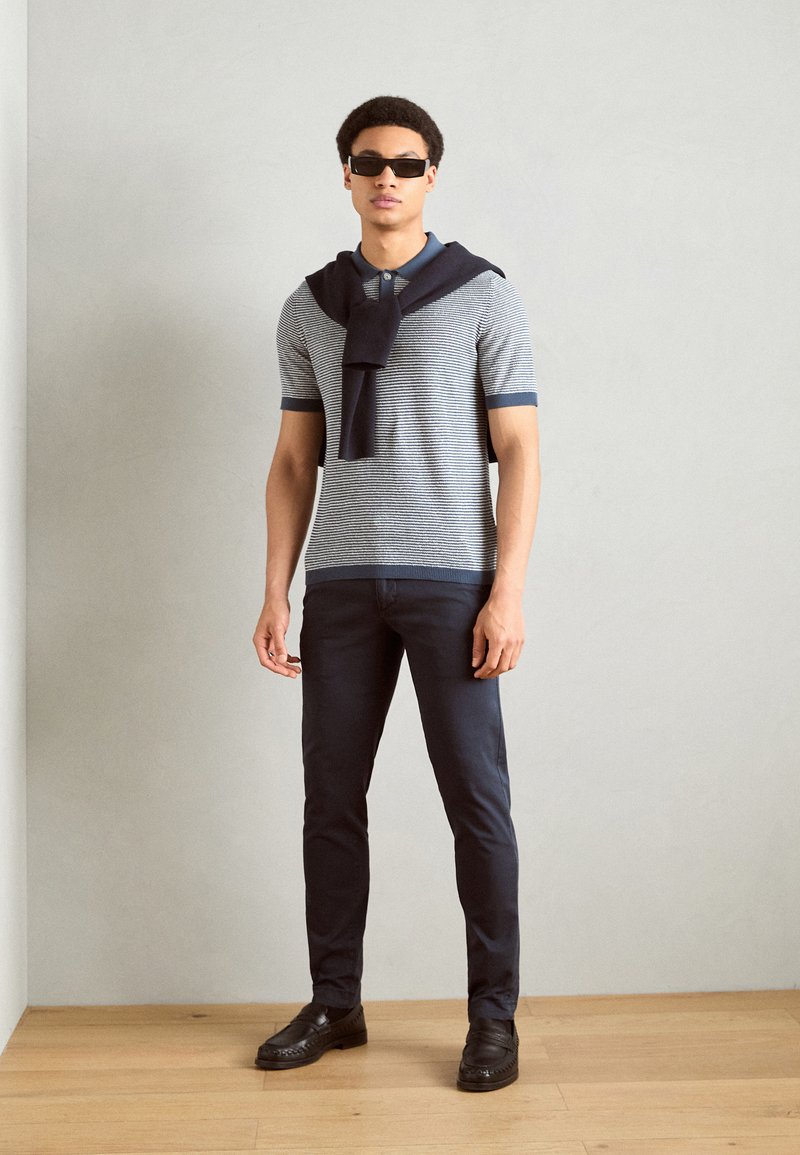 Grau und navy gestreiftes Polohemd mit kurzen Ärmeln, navy Pullover um den Hals gebunden, dunkle Hose und schwarze Slipper. Klare Linien und strukturierte Passform.