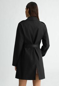 Cappotto lungo nero con vita regolabile, maniche lunghe e spacco posteriore. Tessuto morbido, collo classico e design minimalista.