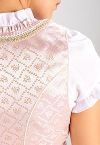 Gros plan d’une personne portant un gilet à motif floral rose et or par-dessus un chemisier blanc à col et manches volantées.