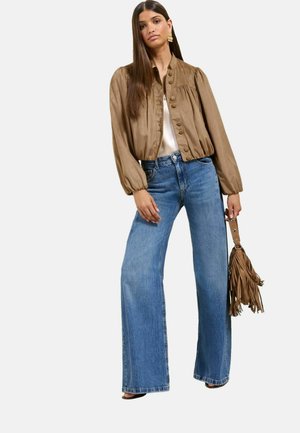 Kobieta w brązowej, zapinanej na guziki satynowej kurtce bomber, białym topie, szerokich niebieskich dżinsach, trzyma brązową torebkę z frędzlami na długim pasku.