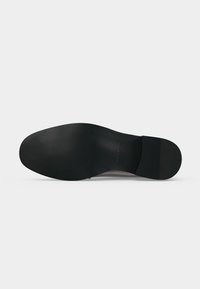 Semelle de chaussure noire vue de dessous, montrant une surface lisse et un léger talon, sur un fond gris clair uni.
