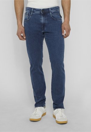 Hombre con vaqueros ajustados azules y zapatillas blancas de pie frente a un fondo liso gris claro.
