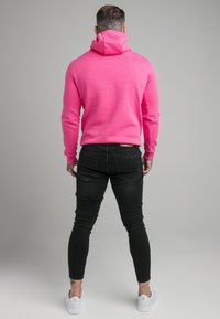 Åtsittande rosa hoodie med hög krage, ribbade ärmslut och mjuk textur, i kombination med svarta slim fit jeans och vita sneakers.