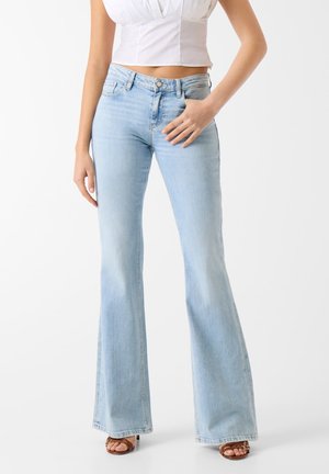 Frau trägt hellblaue hoch taillierte Schlagjeans, weißes, kurz geschnittenes ärmelloses Oberteil und braune Sandalen mit offenem Zehenbereich, steht vor einem weißen Hintergrund.