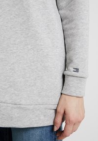 Sweatshirt gris clair avec une finition texturée, présentant un poignet côtelé et un petit accent marine sur la manche. Porté avec un jean en denim bleu.