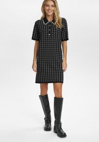 Musta polo mekko, jossa on valkoinen ruutukuvio, lyhyet hihat ja napitus. Yhdistetty polvenkorkuisiin mustiin nahkasaappaisiin.