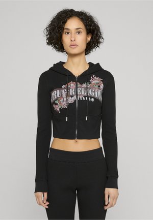 Felpa nera corta con zip e tessuto strutturato, caratterizzata da ricami floreali e coulisse. Abbinata a pantaloni neri coordinati con tessuto strutturato.