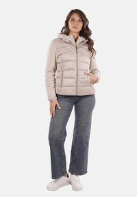 Giacca puffer beige chiaro con cappuccio, chiusura con zip e tasche laterali. Abbinata a jeans blu e sneakers bianche. Texture liscia.