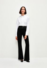 Camicia bianca con colletto, pantaloni neri svasati con cintura a fibbia e scarpe nere. Tessuto liscio e vestibilità su misura, design minimalista.