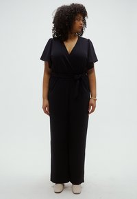 Fekete, rövid ujjú, átlapolt overall puha anyagból. Derékban megkötős övvel és bőszárú fazonnal. Semleges színű cipők.