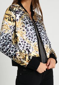 Bomberjack met een glanzend, patroonoppervlak met een zwart, goud en paarse luipaardontwerp, zwarte geribbelde boorden en zoom, ritssluiting.