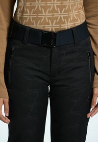 Zwarte gevoerde broek met een subtiel geometrisch patroon, voorzien van ritszakken en een zwarte elastische riem met een glanzende gesp. Bruine langemouwen top.