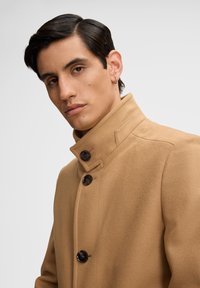 Cappotto in lana color cammello con colletto alto, dotato di tre bottoni scuri e una texture liscia. Il design è sartoriale e aderente.
