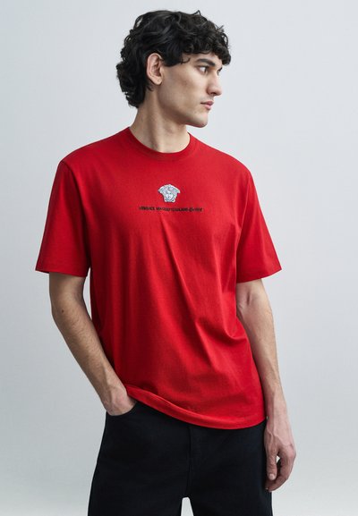 Rotes Baumwoll-T-Shirt mit Rundhalsausschnitt, kurzen Ärmeln und einem schwarzen grafischen Logo mit Medusa-Emblem auf der Vorderseite.
